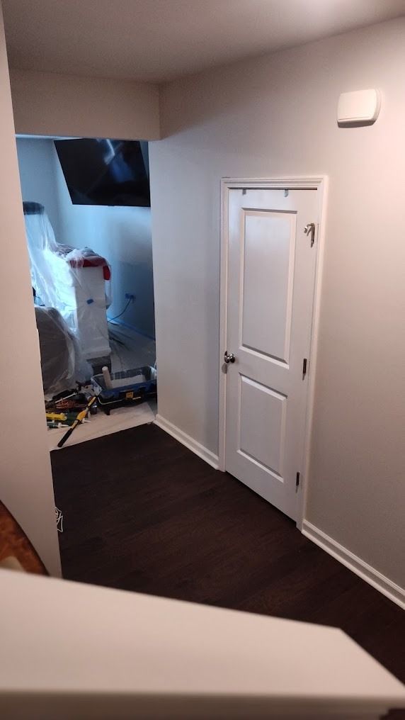 Narrow hallway with dark floor, white door, and a cluttered, partially demolished room at the end