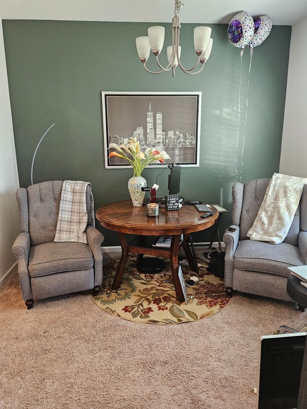 Cozy living room with two gray recliners, round wooden table, green walls, chandelier, and balloons.