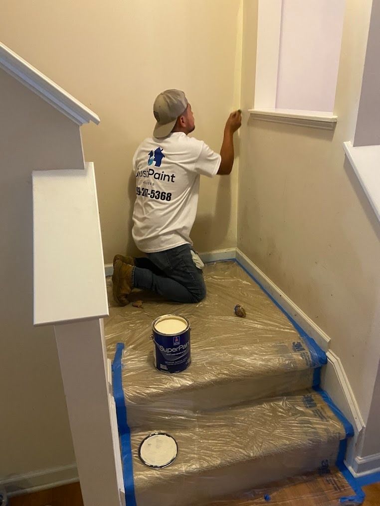 Painter kneeling on stair landing, painting a beige wall with blue tape and a paint can nearby.