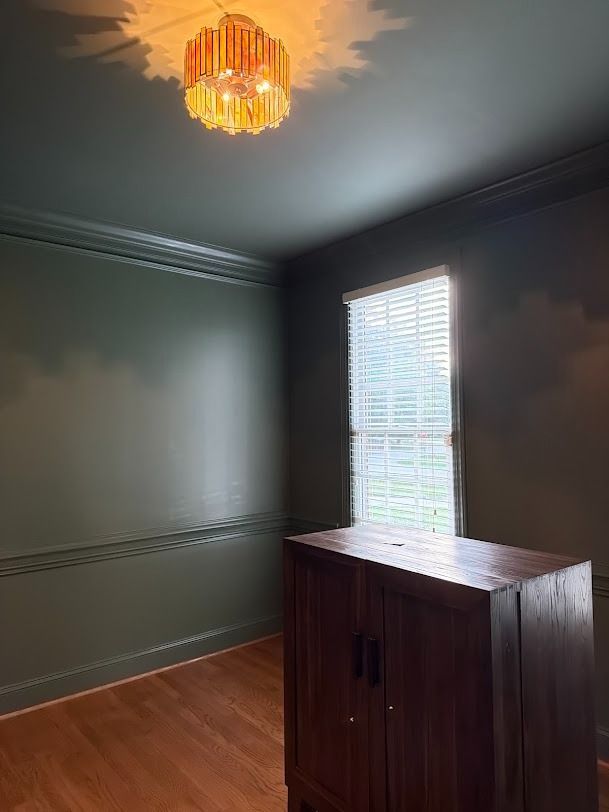 Empty room with dark green walls, wood floor, window blinds, and a wooden cabinet under a ceiling light.