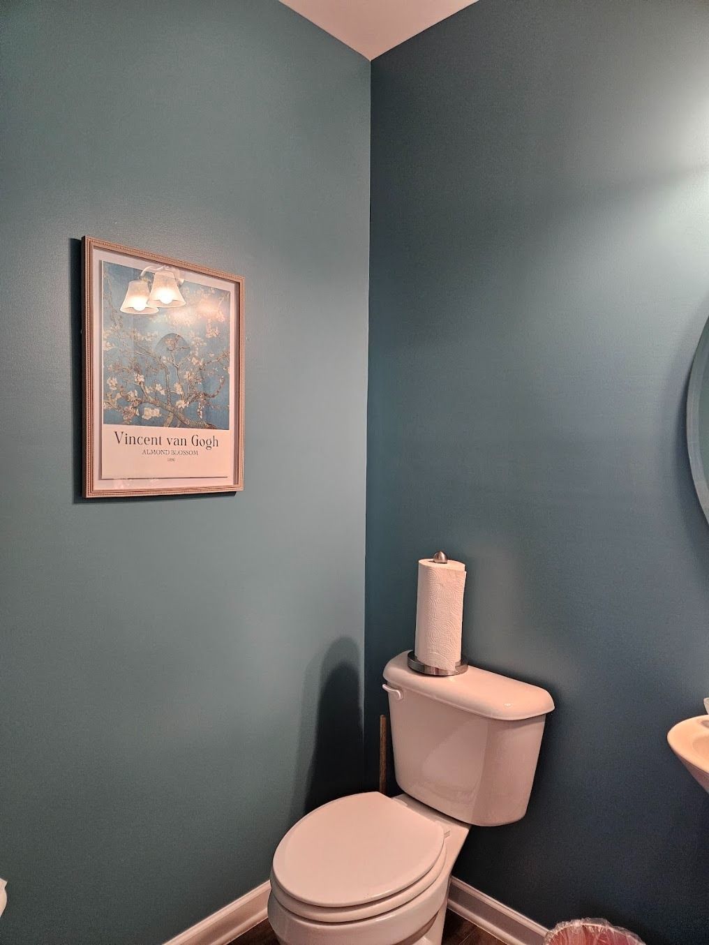 Teal bathroom with a toilet, paper towel roll, framed wall art, and sink edge visible