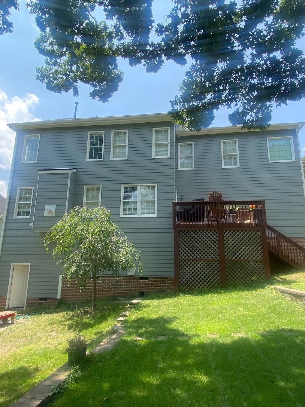 Back view of a blue two-story house with a wooden deck, stairs, and a grassy yard under trees.