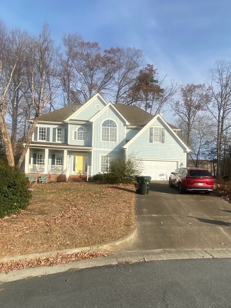 Two-story light-blue suburban house with a driveway, red car, and leafless trees in front