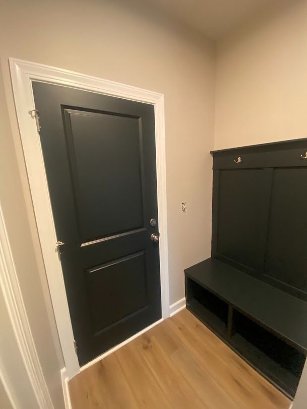 Small entryway with a black door, light wood floor, and black bench with storage hooks.