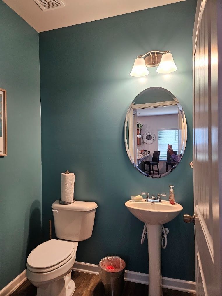 Small teal bathroom with toilet, pedestal sink, oval mirror, wall light, and window reflection.