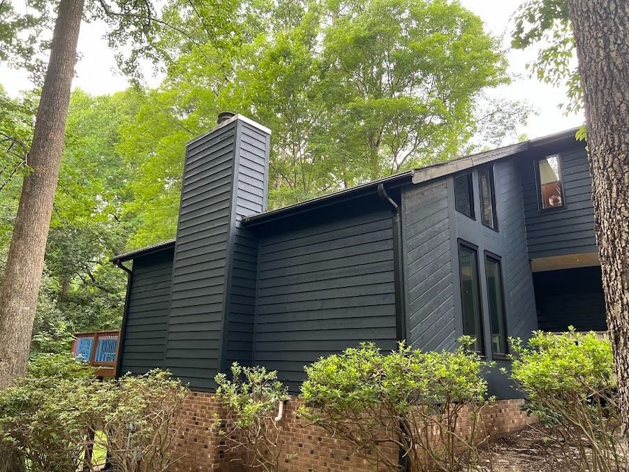 Dark charcoal modern cabin among green trees with tall chimney and shrubs in front