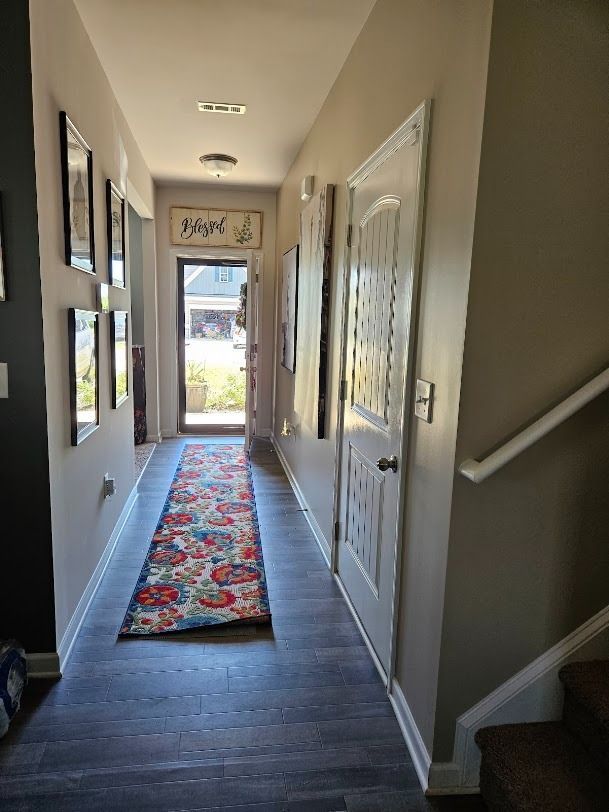 Narrow hallway with dark floor, colorful runner rug, white doors, stairs, and a bright front entry door.