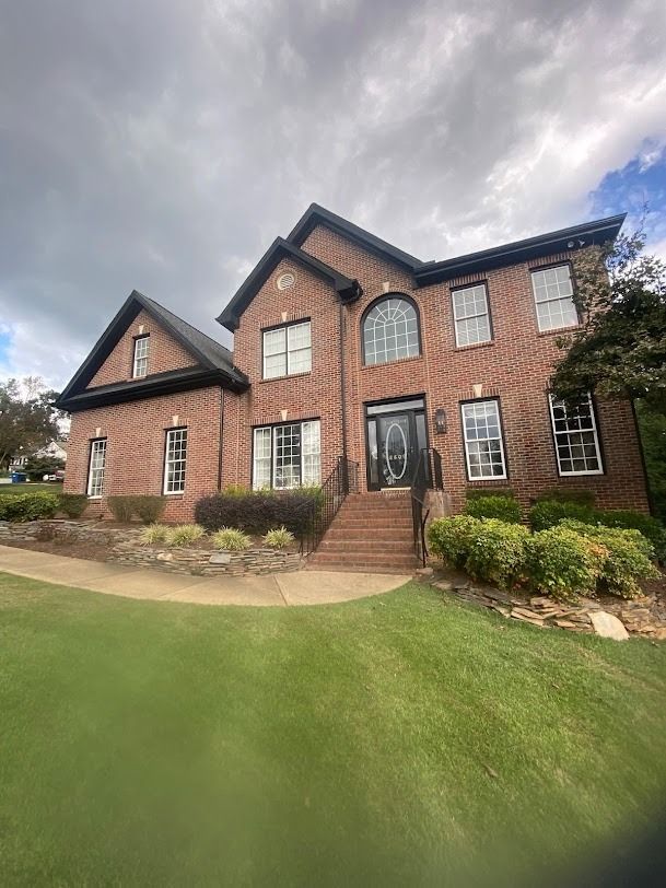 Brick two-story house with gabled roof, manicured lawn, and cloudy sky.