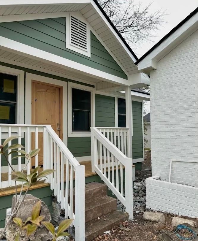 Front porch of a green house with white trim and railings next to a white brick building.