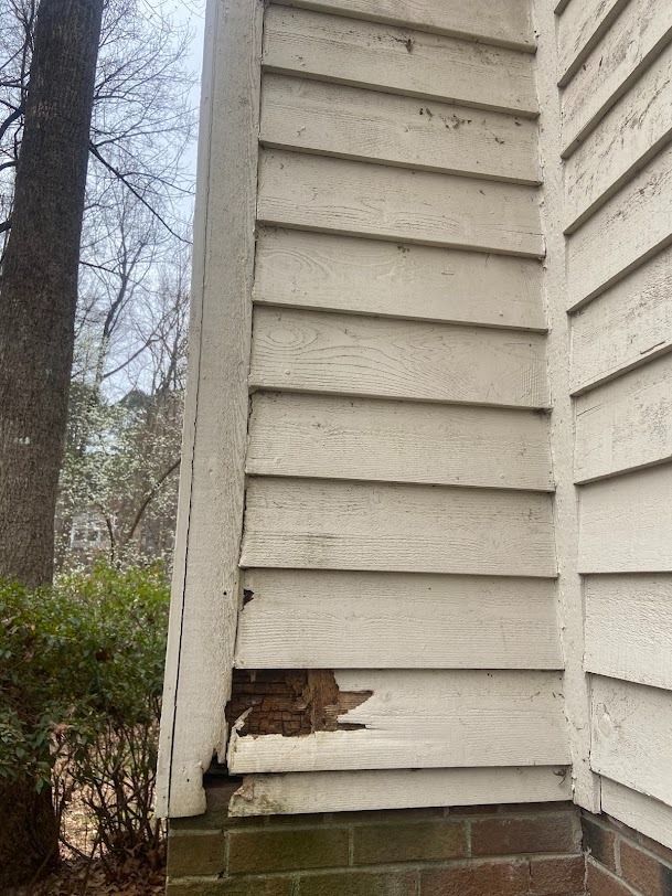 Damaged beige siding on a house corner with exposed wood near the bottom panel