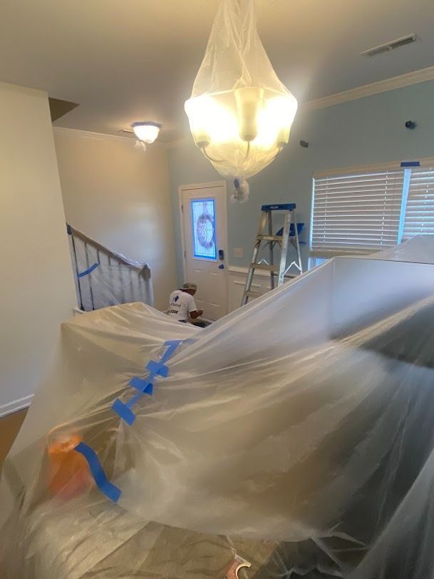 Plastic-covered room with blue tape, ladder, and chandelier during painting or renovation