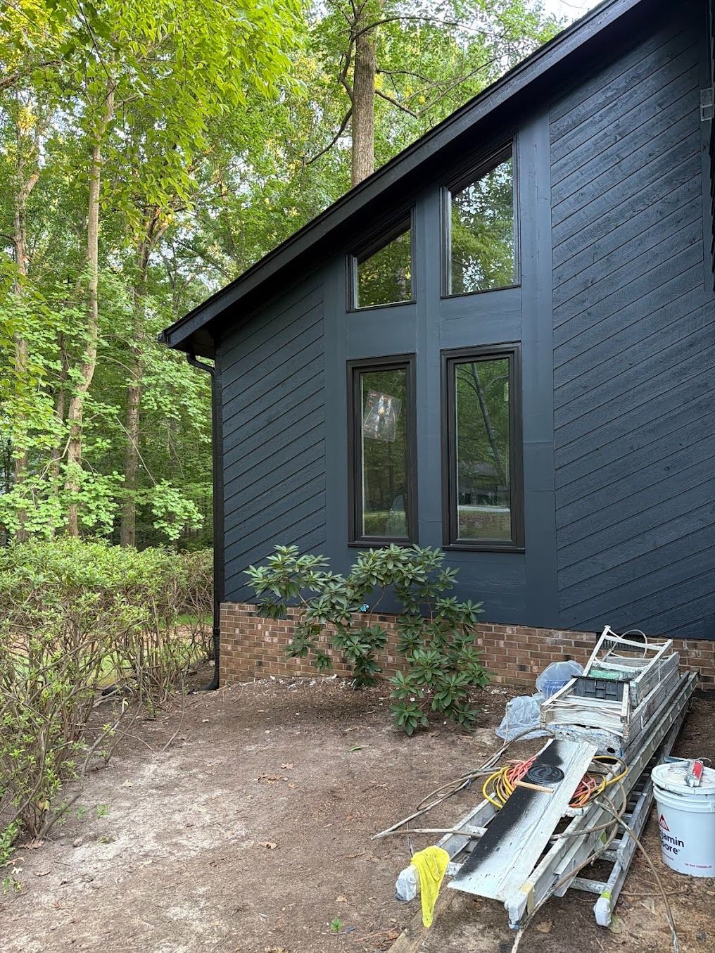 Dark gray cabin exterior with tall windows, surrounded by woods and a pile of ladders and tools nearby