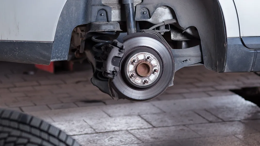 A car with its tire removed, exposing the brake disc and suspension components inside a repair garage.
