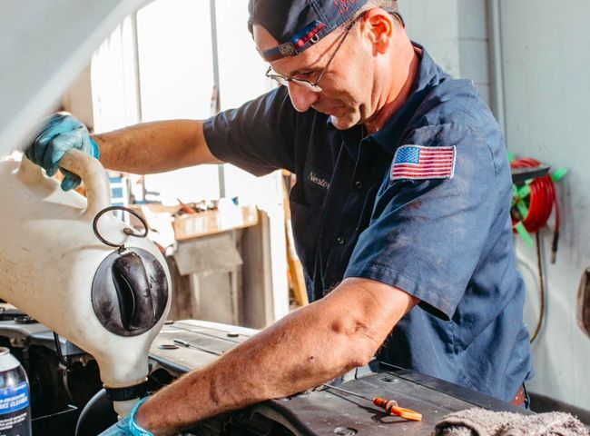 Mechanic pours fluid into car engine. Garage setting, focused expression, blue shirt, gloves, and American flag patch.