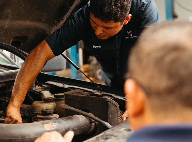 Mechanic working on a car engine, another person watching in an auto repair shop.