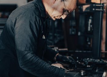 Man in dark work clothes, glasses, and gloves, working at a machine in a workshop.