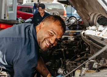 Mechanic smiling while working on a car engine. Another mechanic in the background.