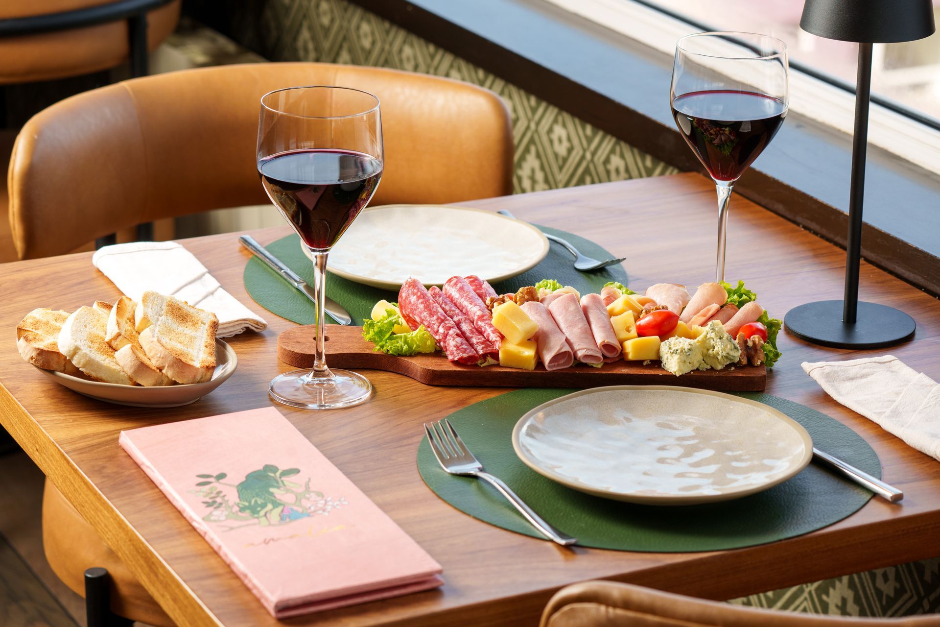 Mesa de restaurante puesta con vino, tabla de embutidos, pan y platos sobre manteles individuales verdes.