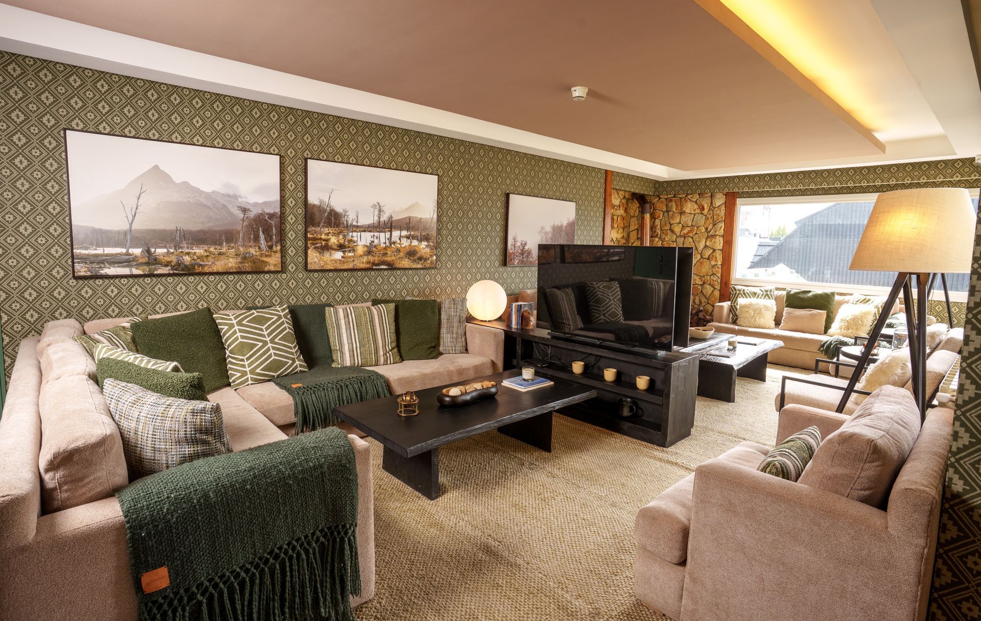 Acogedora sala de estar con sofás beige, mesa de centro oscura, cuadros en la pared, televisor y grandes ventanales con iluminación cálida.