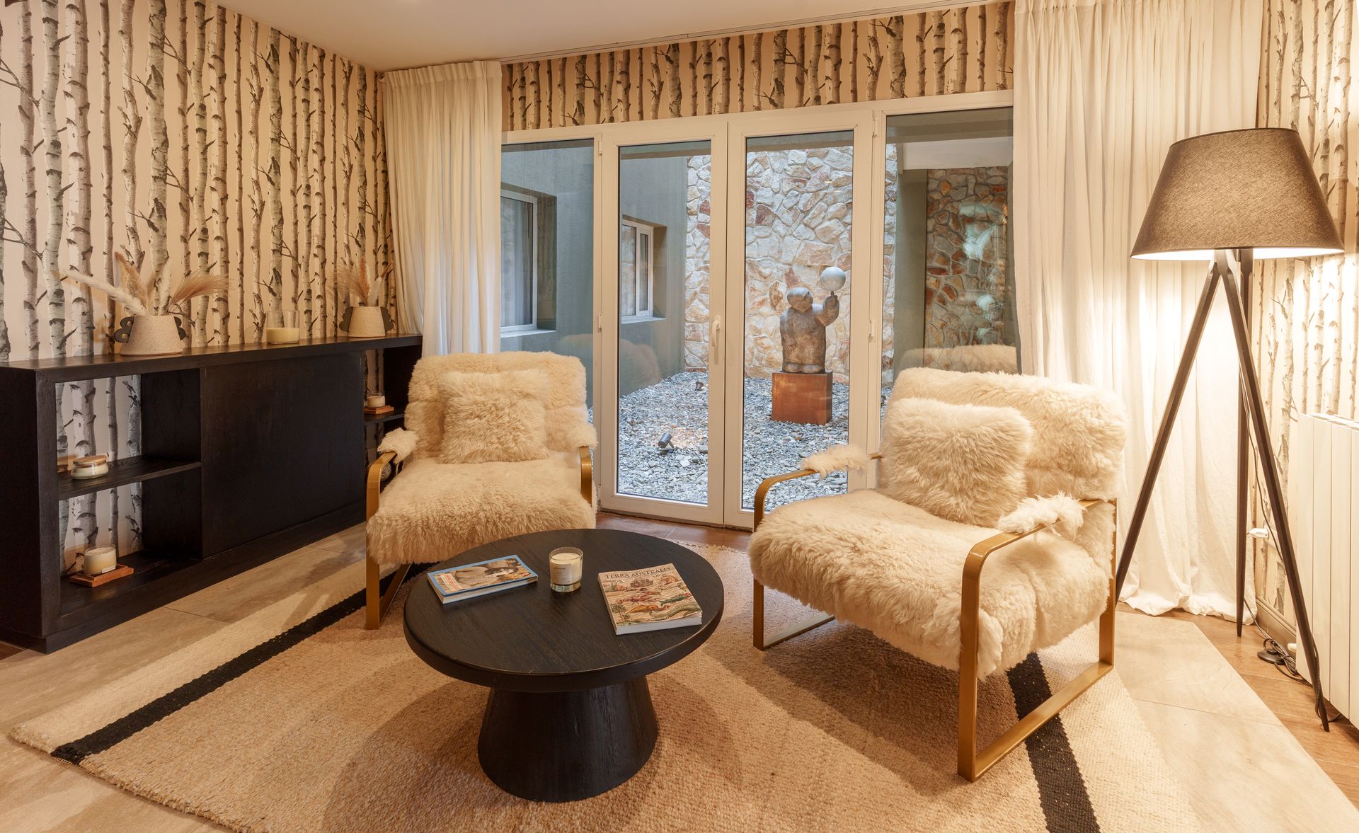 Acogedora sala de estar con dos sillones color crema, mesa de centro redonda negra, lámpara y puertas corredizas de cristal.