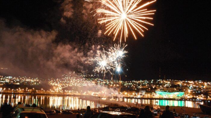Fuegos artificiales sobre una ciudad y un paseo marítimo brillantemente iluminados por la noche.