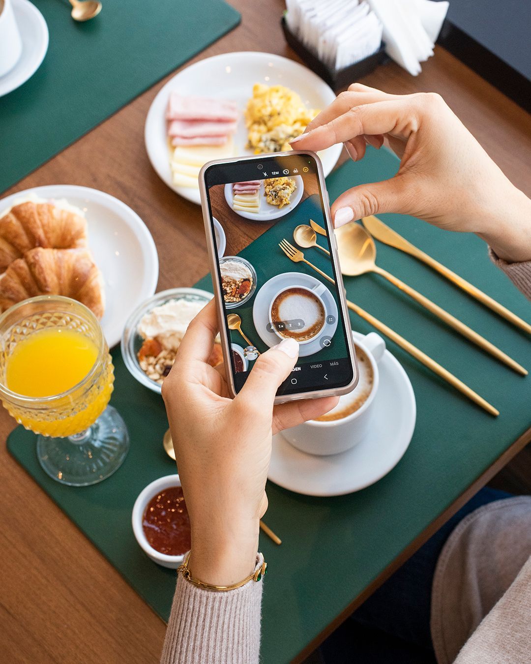 Manos fotografiando una mesa de desayuno con café, croissants, zumo de naranja y palillos chinos sobre un mantel individual verde.