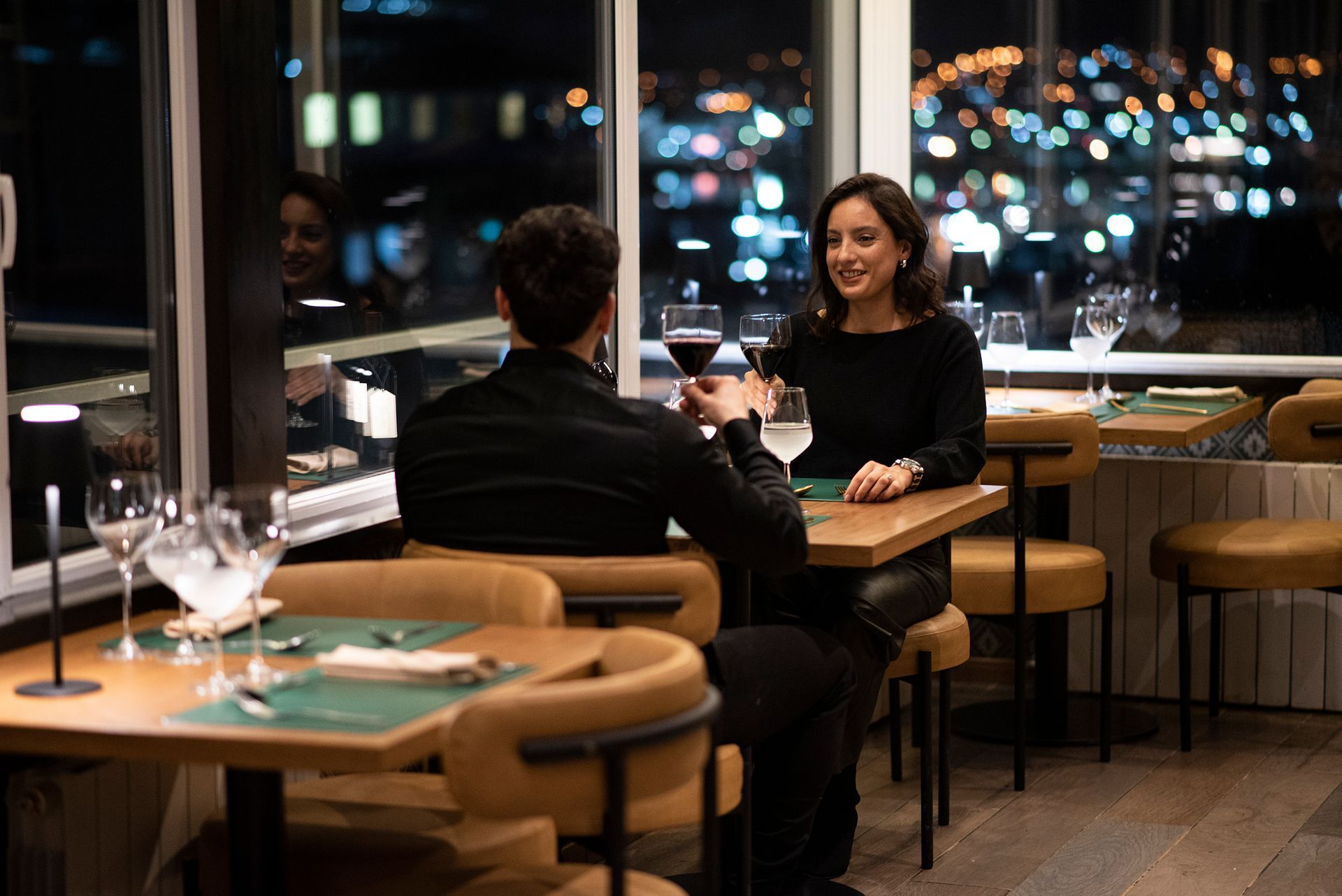 Dos personas charlando en la mesa de un restaurante junto a una ventana por la noche, con las luces de la ciudad iluminadas desde fuera.