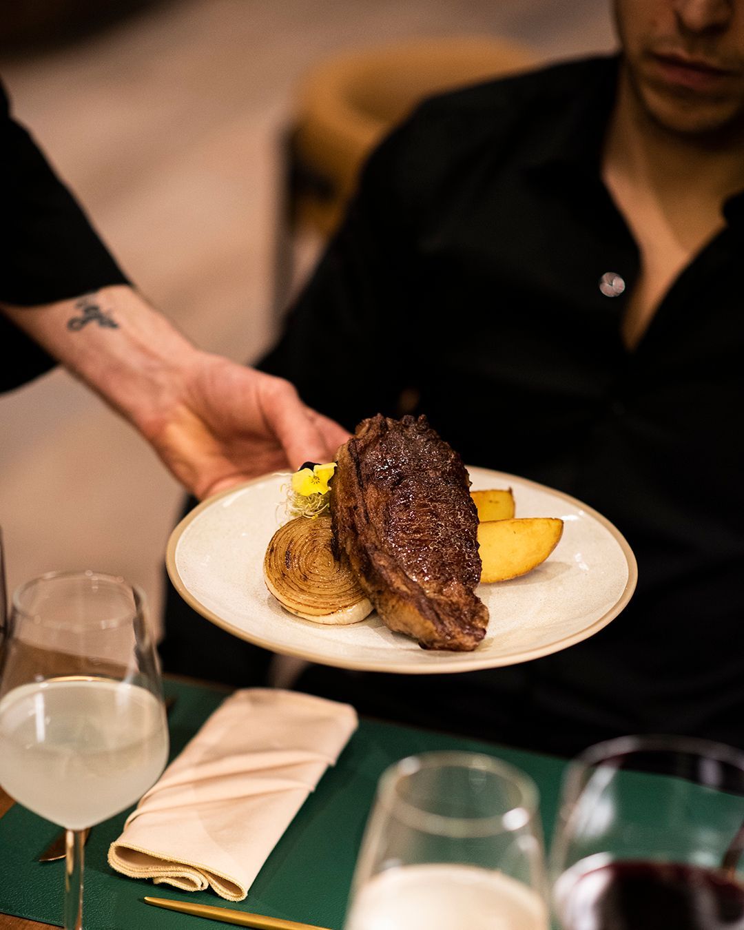 Un camarero coloca un plato de bistec en la mesa junto a una copa de vino blanco.