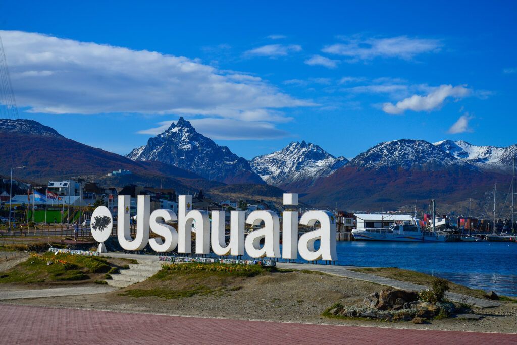 Letrero de Ushuaia junto al paseo marítimo con montañas nevadas y cielo azul al fondo.