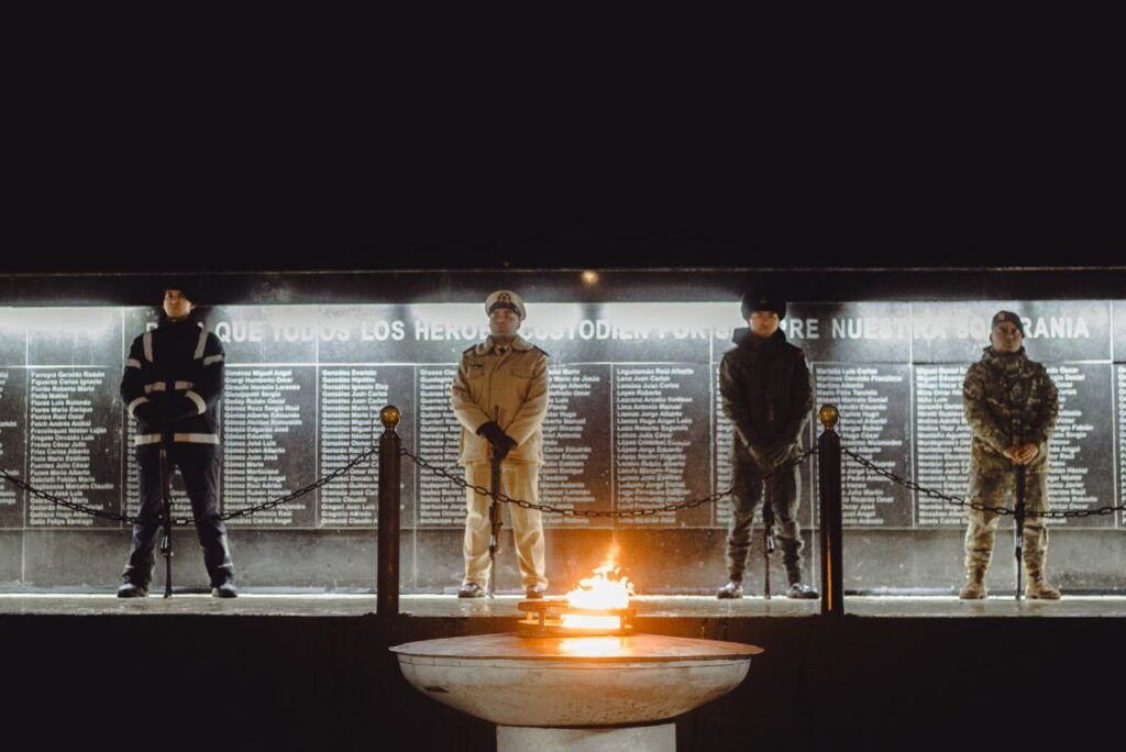 Cuatro personas permanecen de pie frente a un muro conmemorativo iluminado por llamas, en un ambiente de ceremonia sombría.