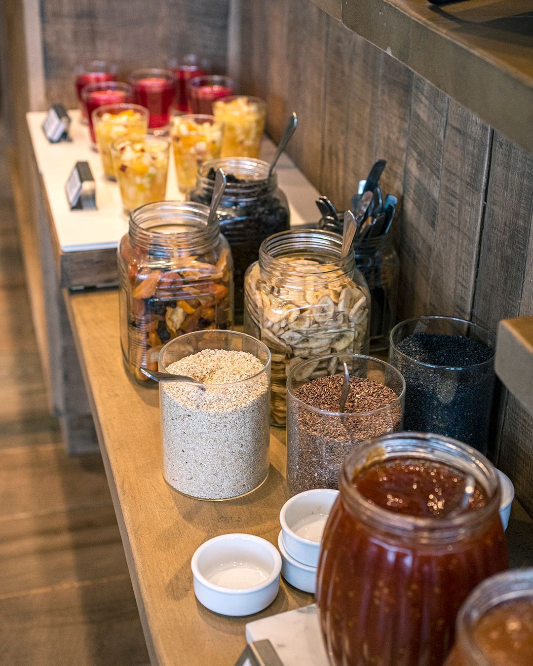 Frascos de cereales, frutos secos y condimentos alineados en un mostrador de buffet con pequeños cuencos y tazas.