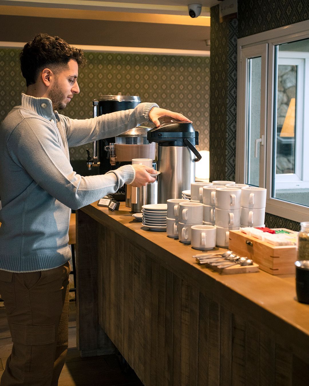 Una persona sirve café de un dispensador en una barra de desayuno con tazas blancas y pasteles.
