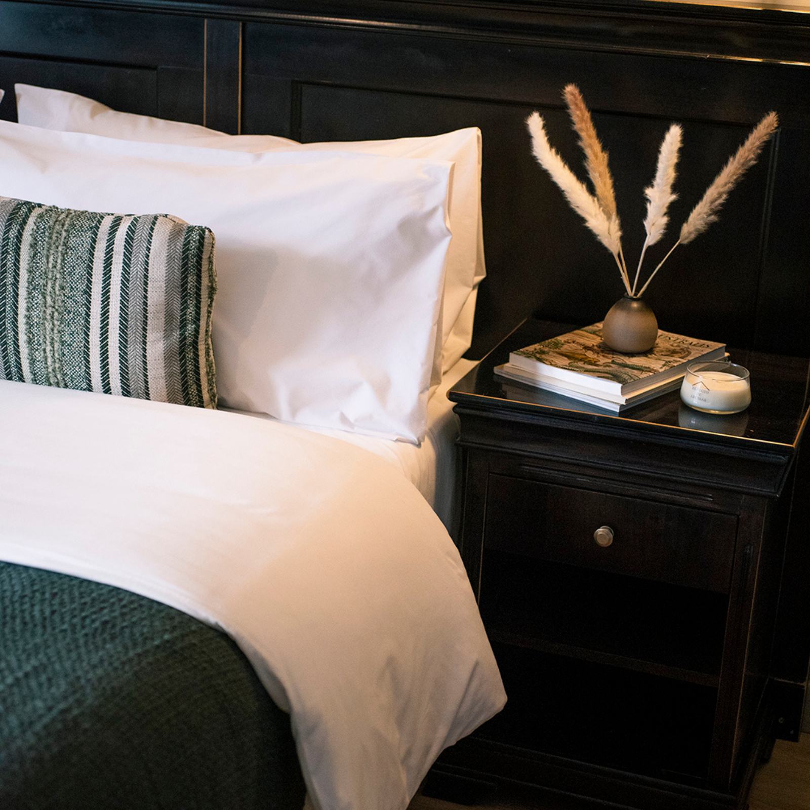 Cama negra con sábanas blancas, almohada a rayas y mesita de noche con libros y hierba de la pampa seca.