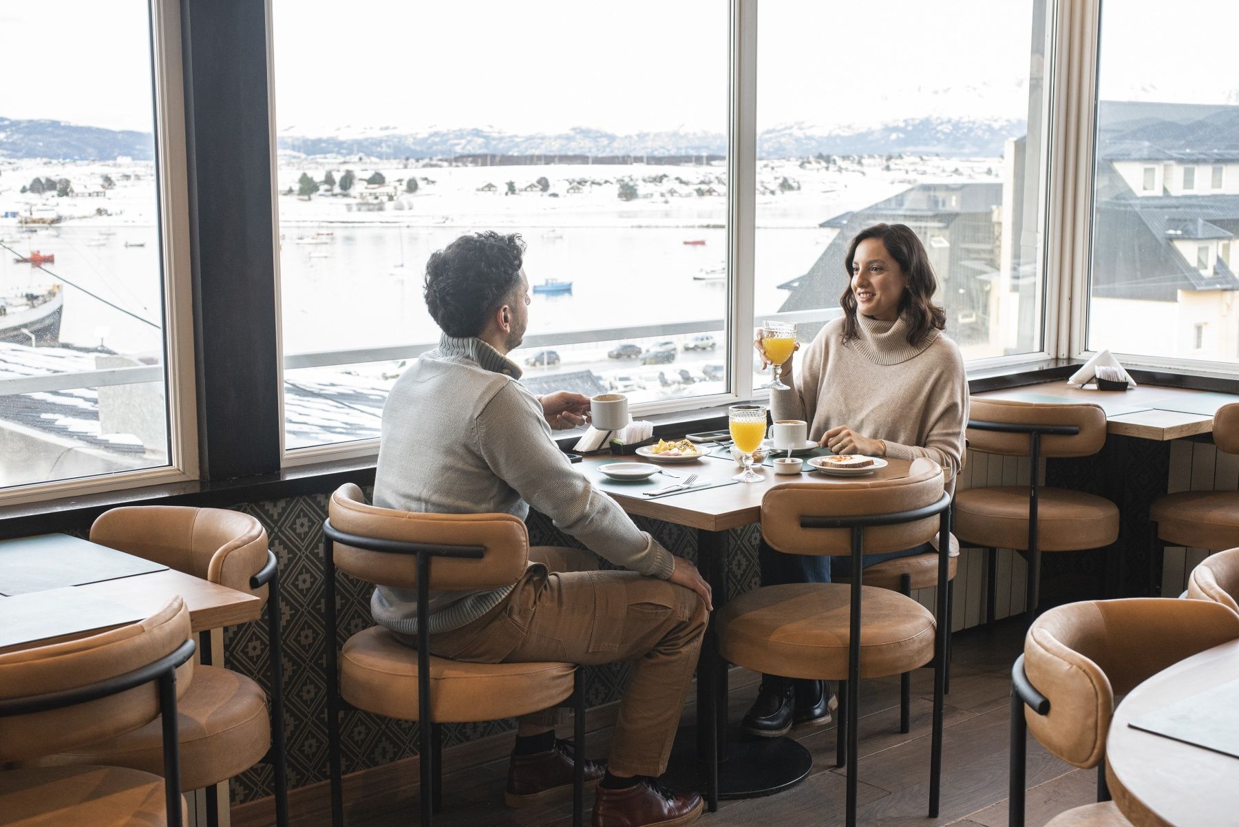 Dos personas charlando en una mesa de café junto a unos grandes ventanales con vistas a un puerto nevado.