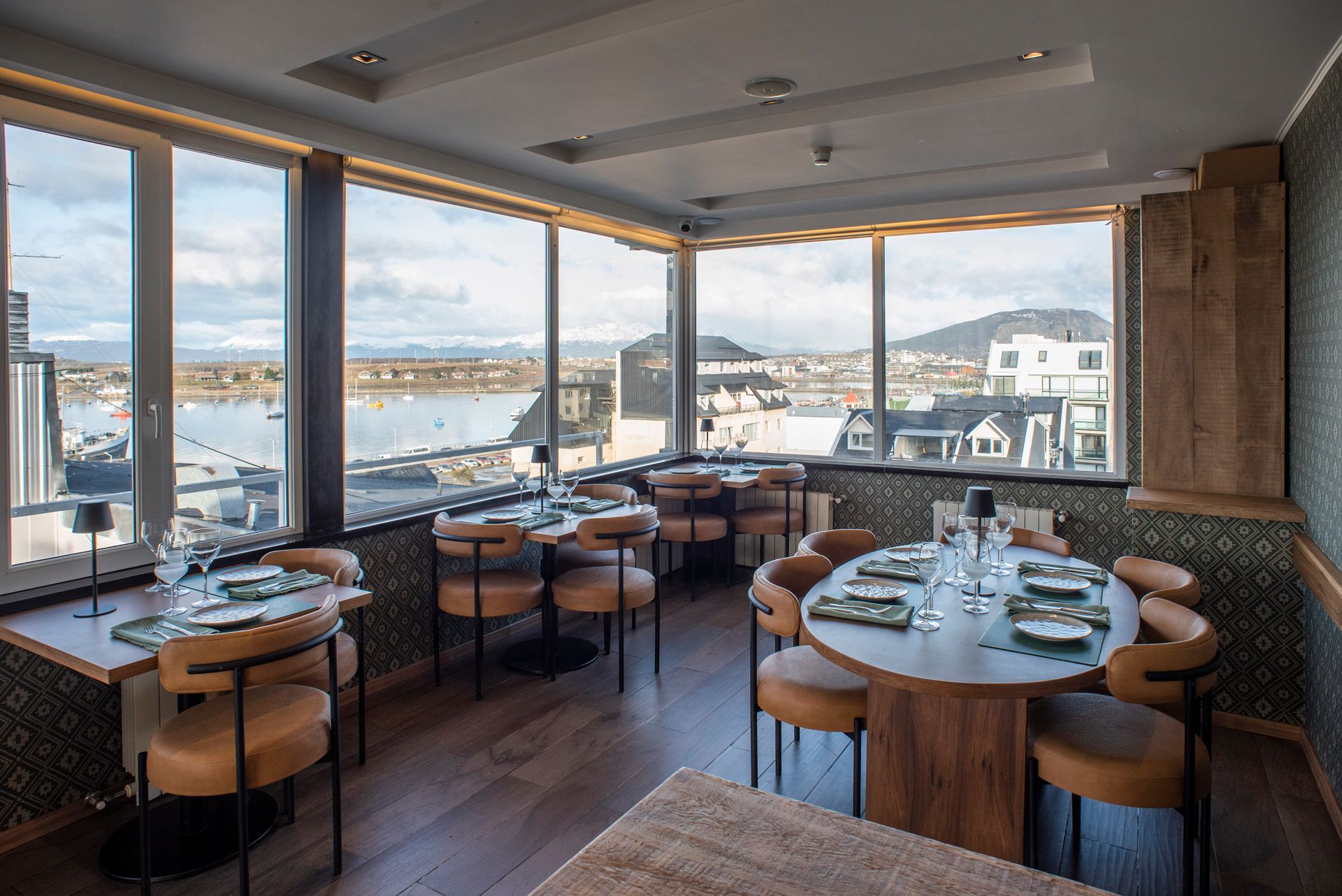 Luminoso comedor de restaurante con mesas redondas y grandes ventanales con vistas al agua y a la ciudad.