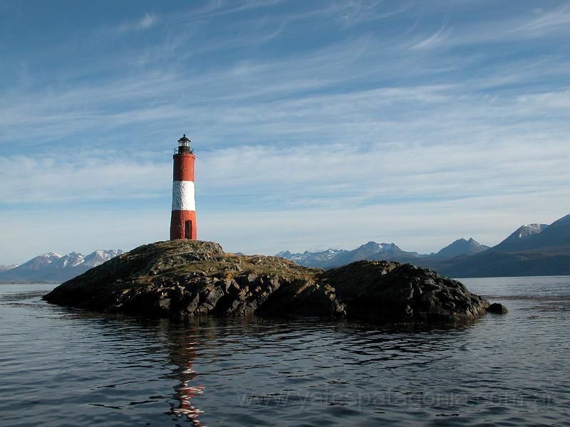 Faro rojo y blanco en una isla rocosa de aguas tranquilas con montañas al fondo.