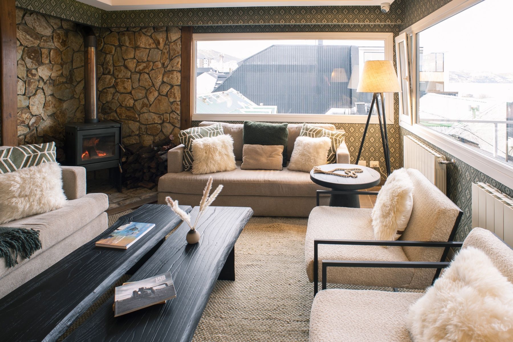 Acogedor salón con paredes de piedra, sofás beige, mesa de centro, chimenea y gran ventanal con vistas a las montañas nevadas.