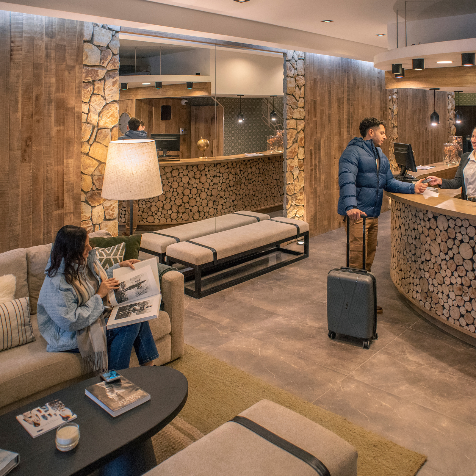 Vestíbulo del hotel con un huésped sentado leyendo, un viajero en la recepción y una cálida decoración de madera y piedra.