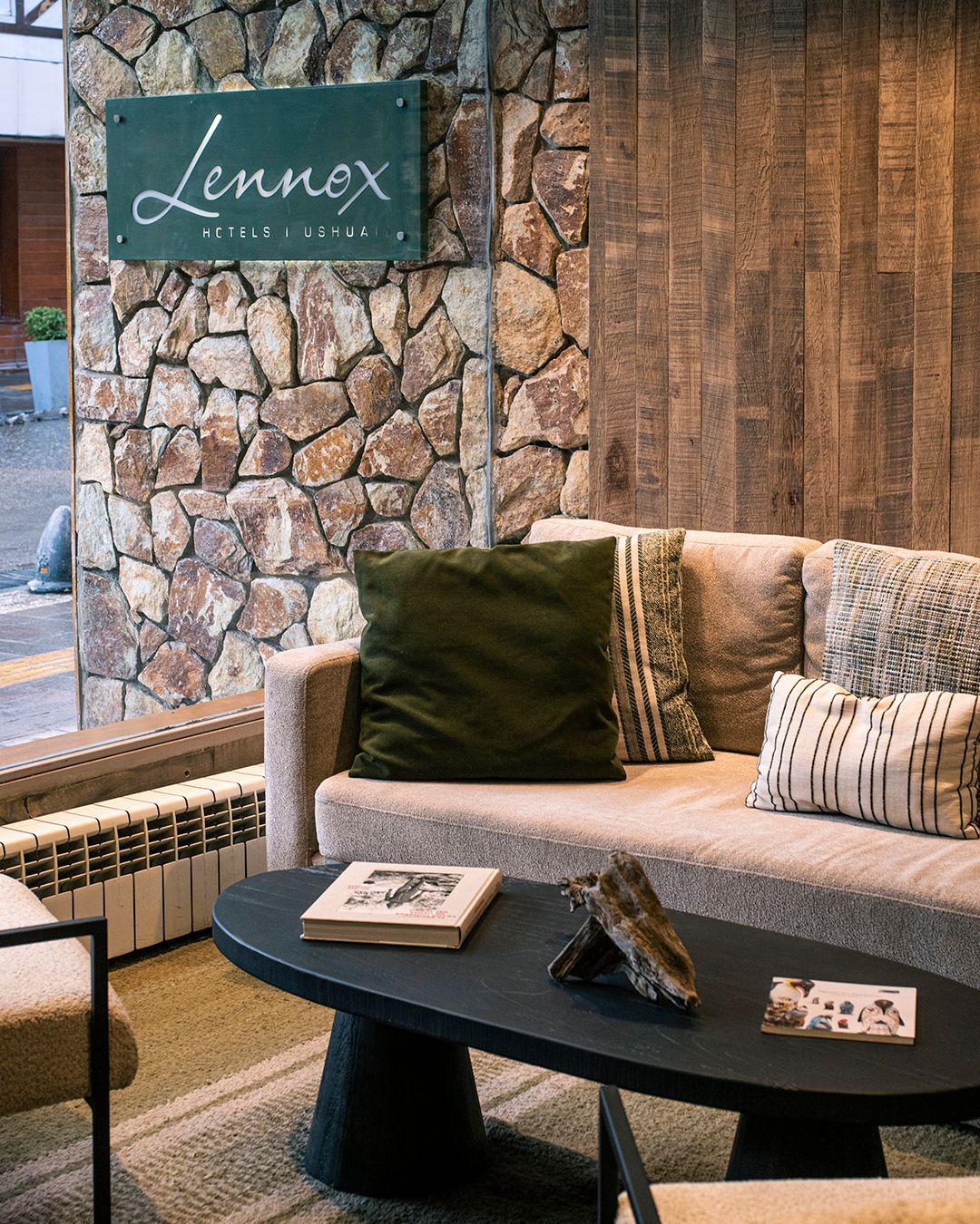 Acogedor salón con pared de piedra, letrero de Lennex, sofá gris, mesa de centro negra y cálidos paneles de madera.