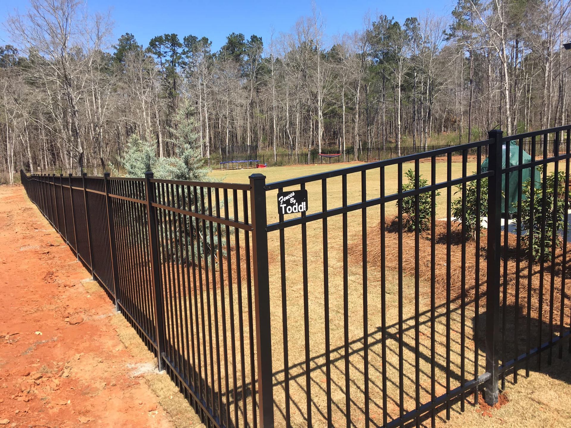 Black metal fence beside a dirt path in a wooded area on a clear day