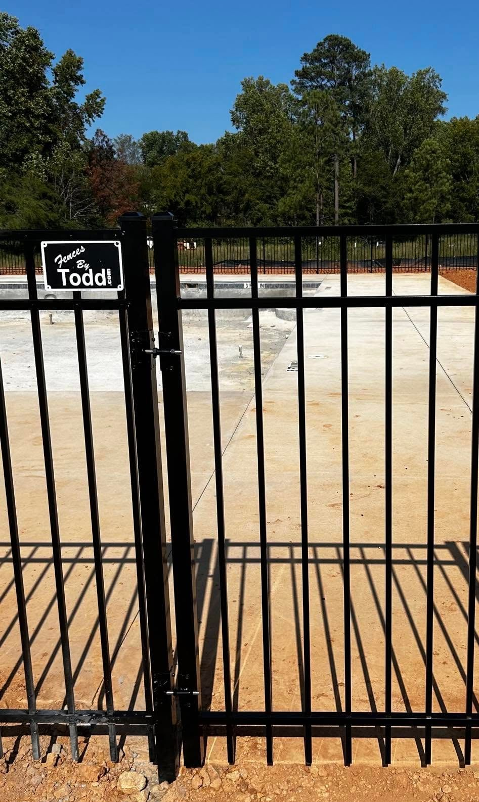 Black metal gate with a small “The Todd” sign, opening onto a sandy yard with trees in the background.