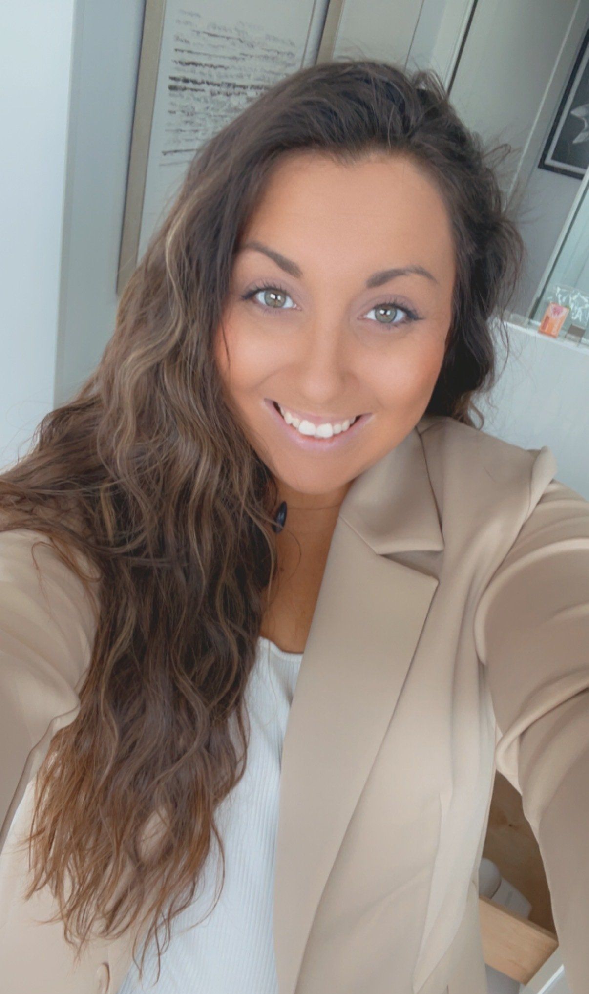 A woman is taking a selfie with her phone while wearing a tan jacket.