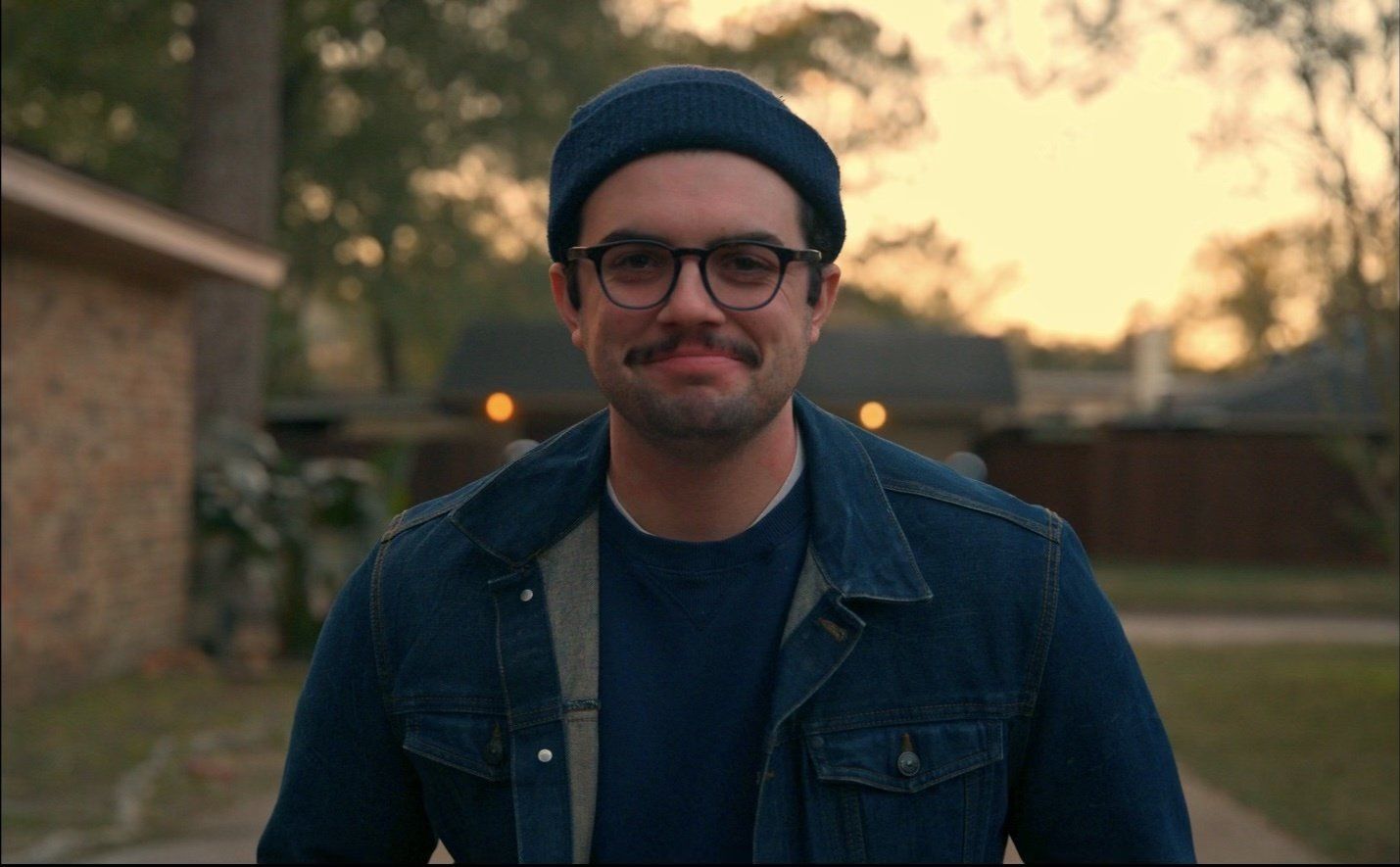 A man wearing a hat and glasses is standing in front of a house.