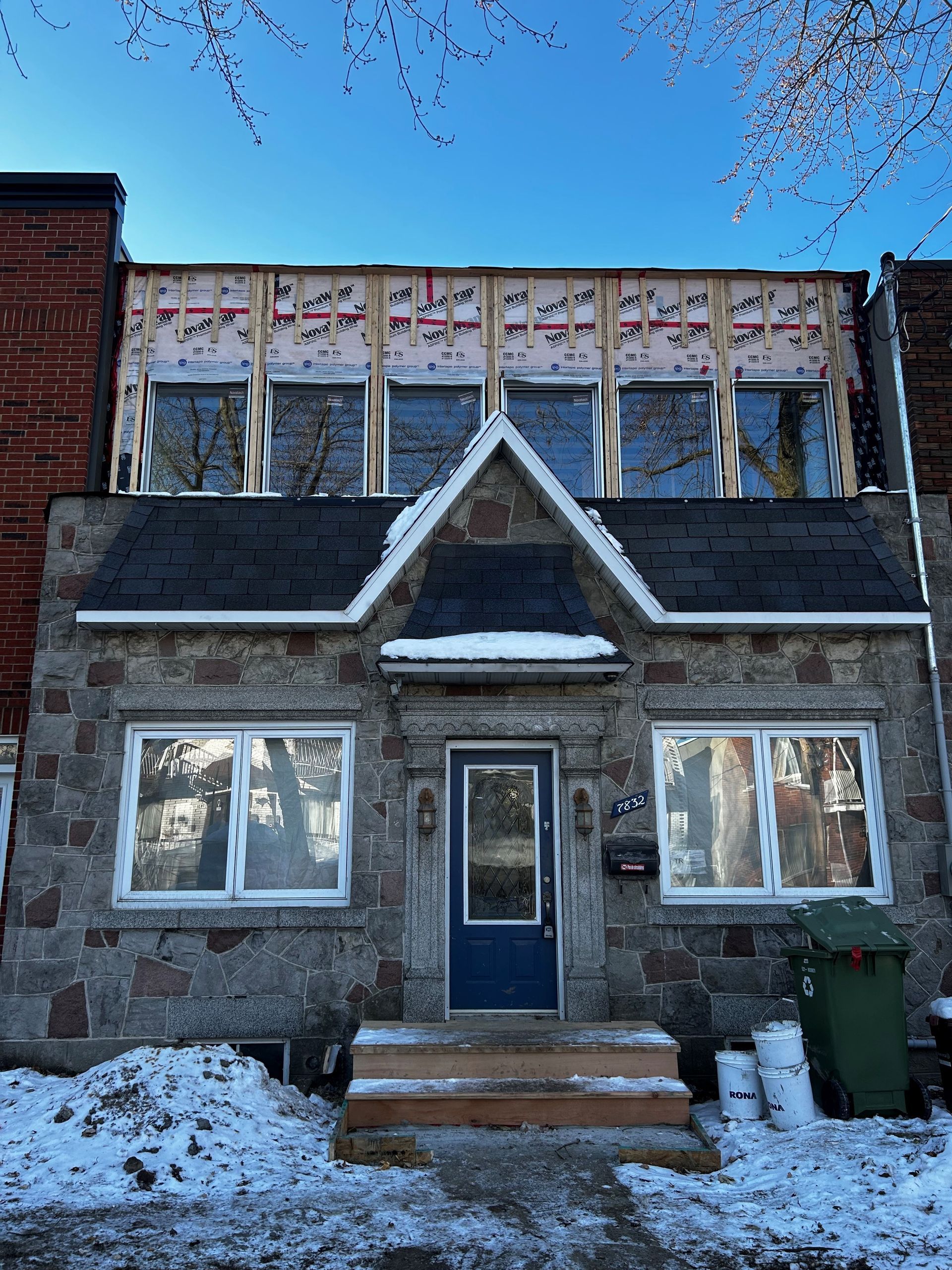 Une maison en pierre avec une porte bleue et un toit noir est recouverte de neige.