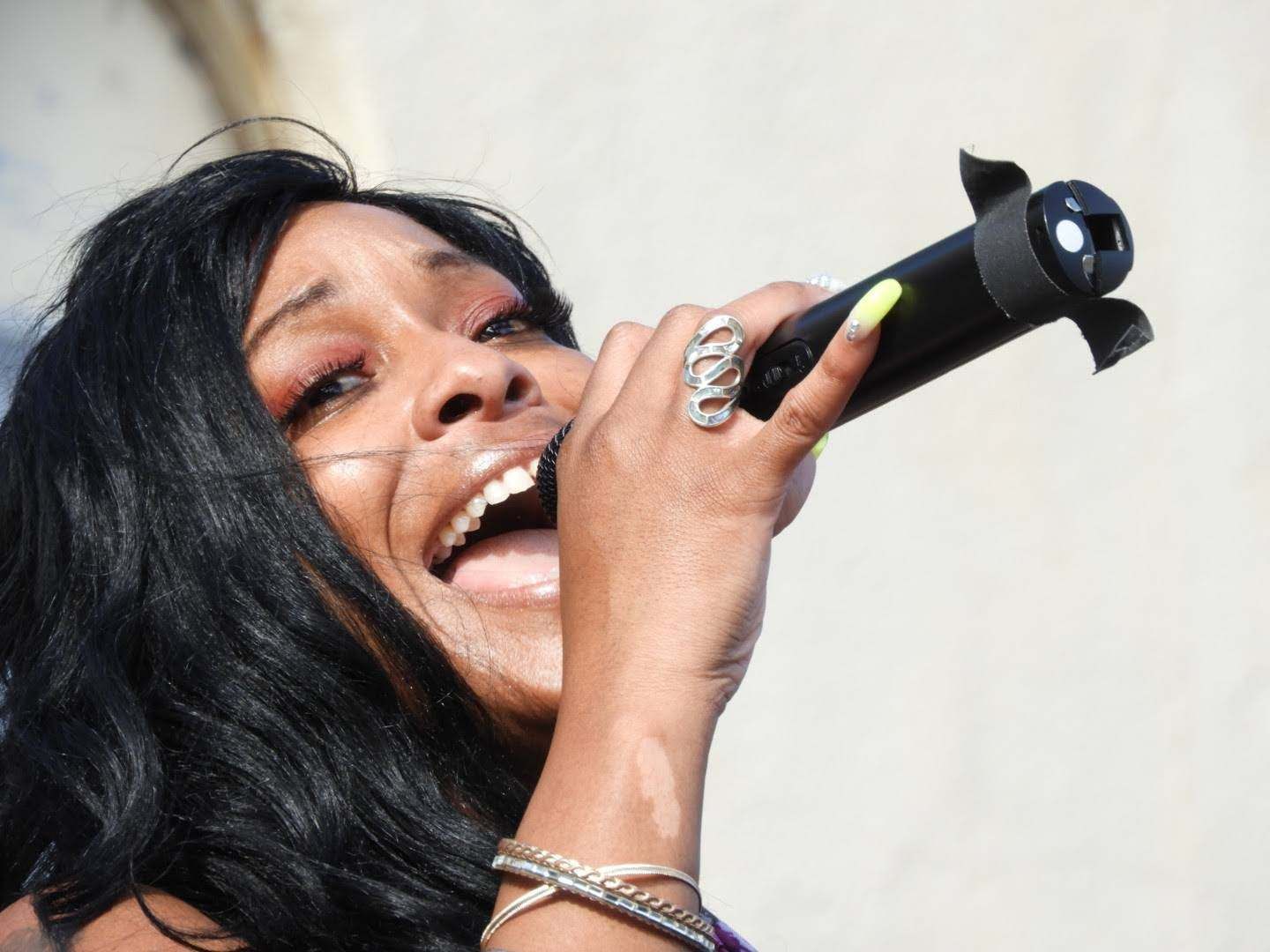 Singer performing into a handheld microphone, smiling, with long black hair and silver jewelry.