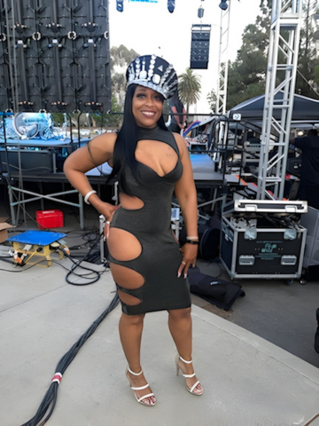 Woman posing on an outdoor stage in a black cutout dress, striped hat, and white heels.