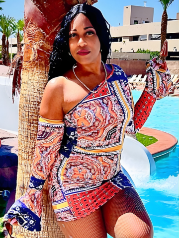 Person in a patterned dress posing by a pool beside a palm tree in bright sunlight