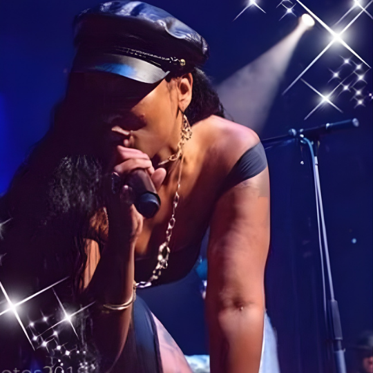 Singer performing onstage under blue lights, leaning into a microphone in a black cap and chain necklace