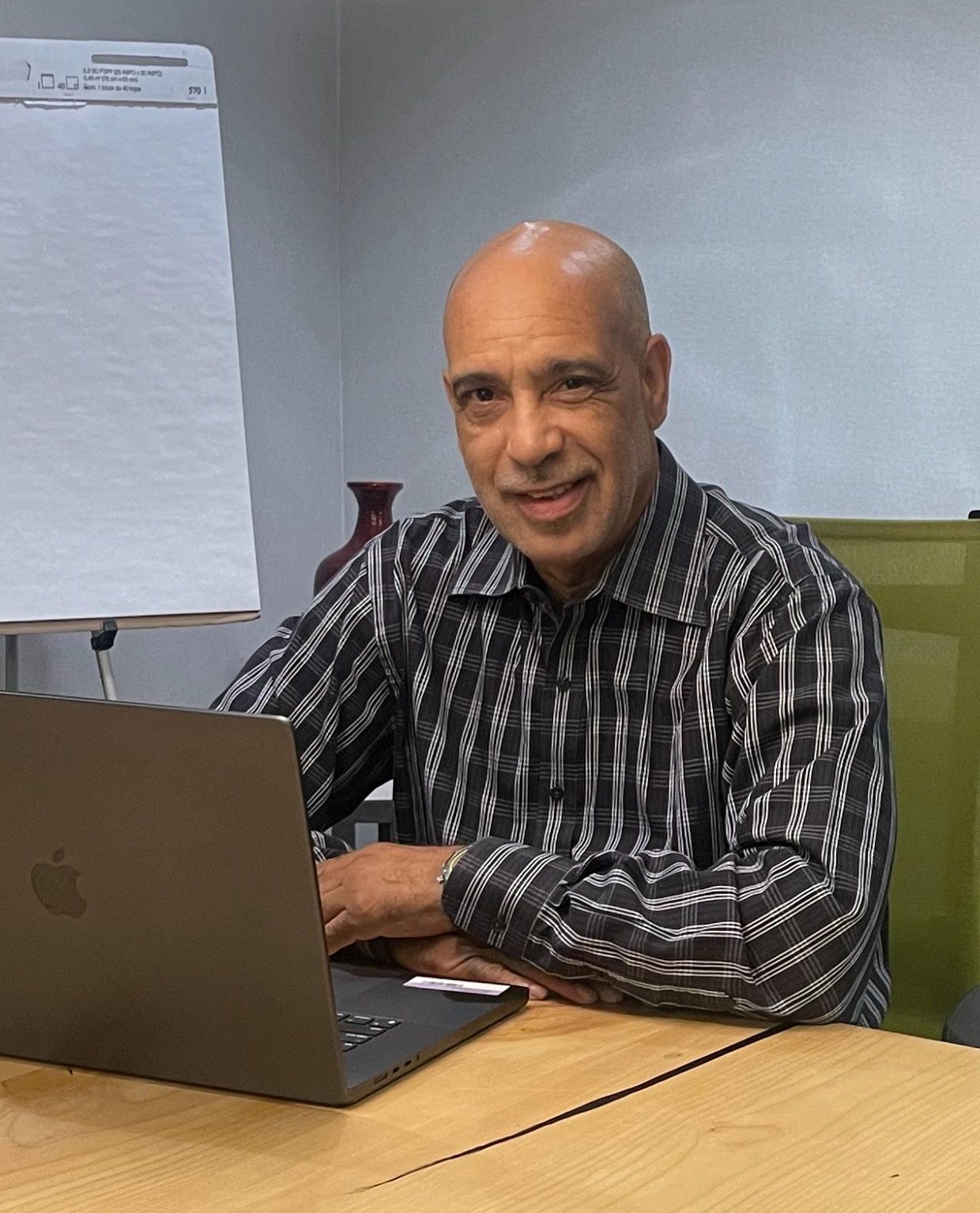 Man sitting at a desk using a laptop in an office, with a whiteboard in the background.
