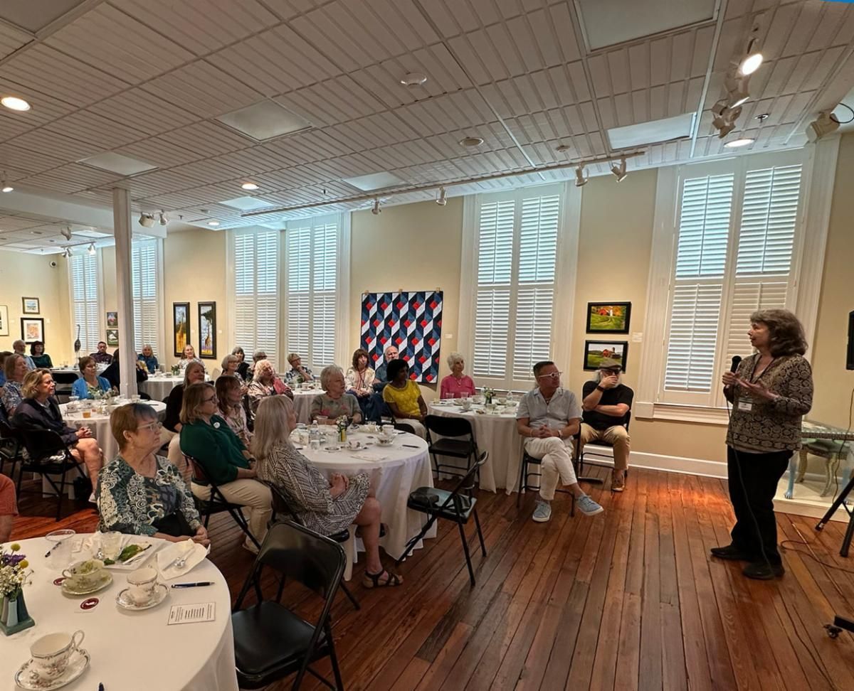 Speaker addressing seated audience in a bright dining room with white tables and hardwood floors